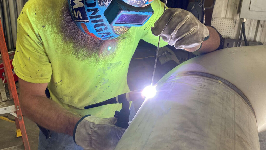 Welder in helmet welding a large metal piece in a workshop setting in landscape orientaion